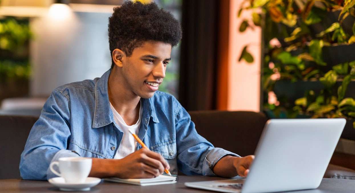 Young man writing notes while using a laptop in a cozy café setting, illustrating effective blogging and content creation strategies.