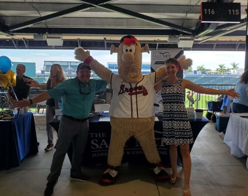 Richard and Wendy Burnham posing with a Braves mascot at The Salt Net marketing booth during a spring training event, showcasing their agency's community engagement and promotional efforts.
