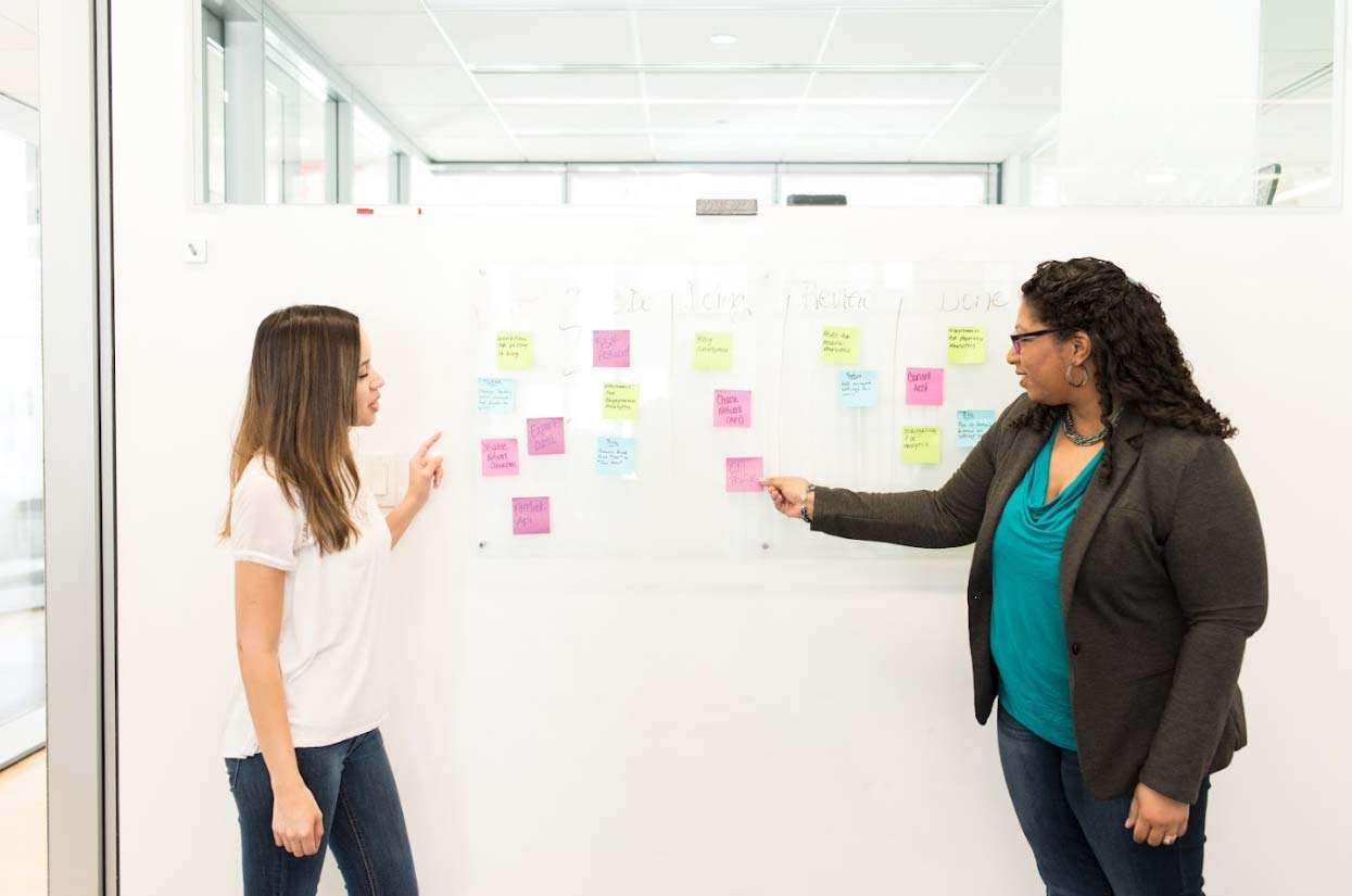 Two women collaborating in an office setting, discussing ideas while pointing at a whiteboard filled with colorful sticky notes, representing brainstorming and strategy development for business growth.