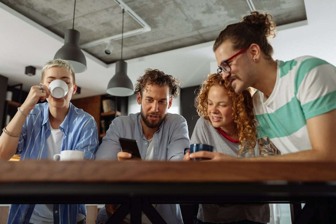 Group of four young adults engaged in discussion over mobile device, coffee cups on table, modern workspace setting, reflecting collaboration and digital interaction related to Salt Net's retargeting solutions.