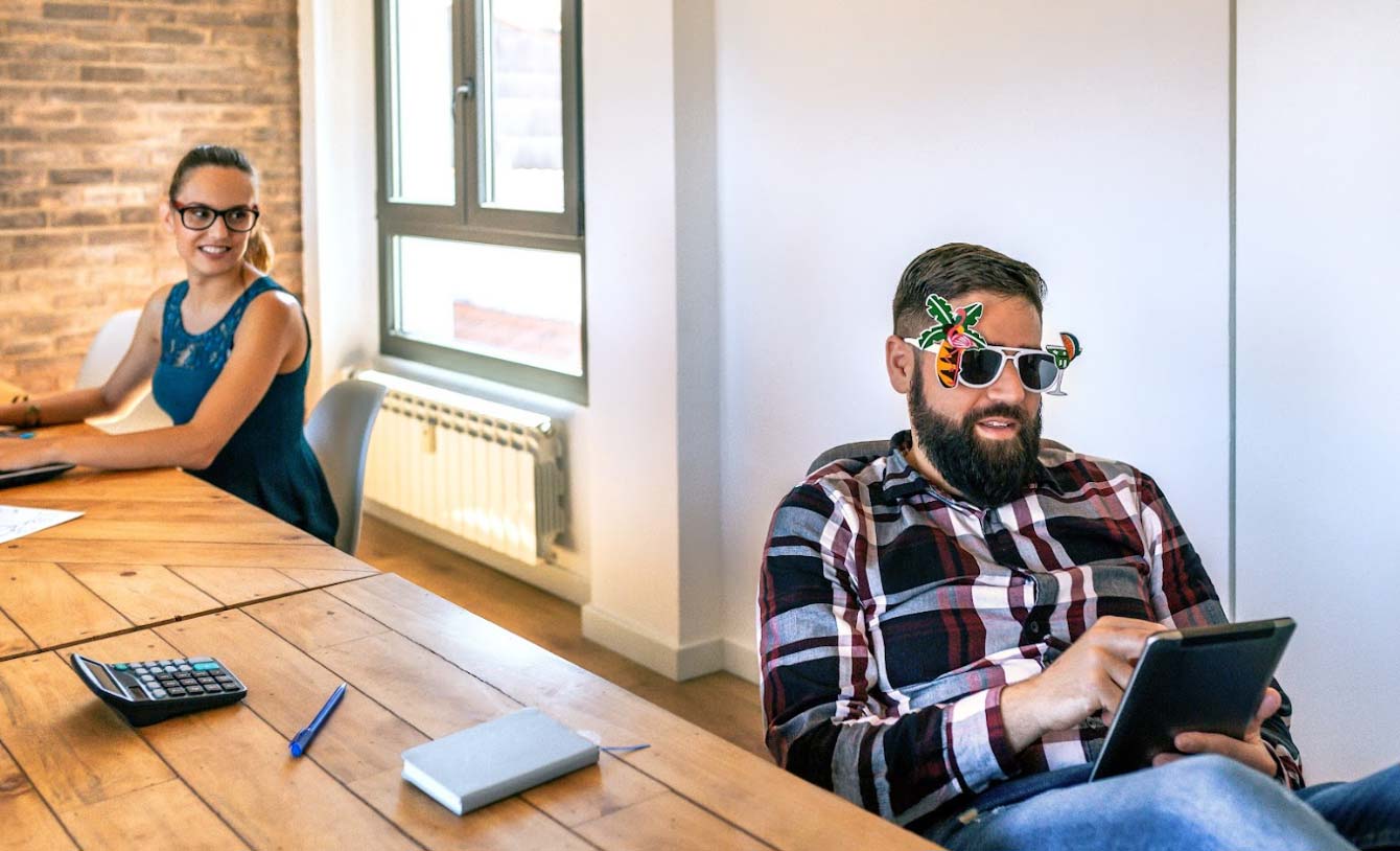 Two professionals in a modern office setting, one woman in a blue dress smiling while working on a laptop, and a man wearing playful sunglasses with tropical designs, using a tablet, emphasizing a creative and engaging work environment.