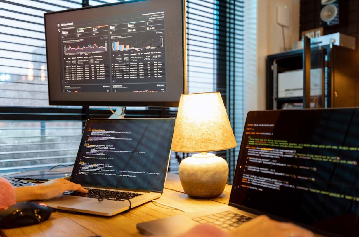 Person working on laptop with coding interface, desktop monitor displaying data analytics dashboard, and lamp illuminating workspace, highlighting web hosting and maintenance services.