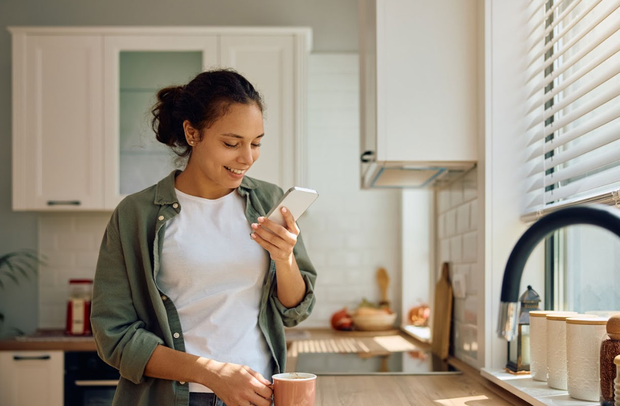 Woman smiling while looking at smartphone in modern kitchen, reflecting customer engagement and digital interaction for AI search optimization strategies.